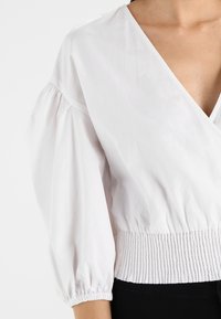 Vit omslagstopp med puffärmar, elastisk midja och djup V-ringning. Slät textur, figursydd i midjan och avslappnad design.