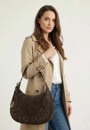 Vrouw in een beige trenchcoat en blauwe spijkerbroek, die een grote donkerbruine schoudertas met studs en een gouden bedel vasthoudt.