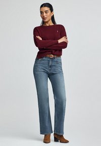Bordeauxfarbener Pullover, gerade geschnittene blaue Jeans und braune Stiefeletten mit Absatz. Der Pullover hat einen Rundhalsausschnitt und ein gesticktes Logo. Lässiges, bequemes Outfit.
