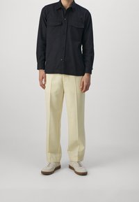 Black, button-up shirt with two chest pockets; paired with yellow, wide-leg trousers; finished with white sneakers with textured soles.