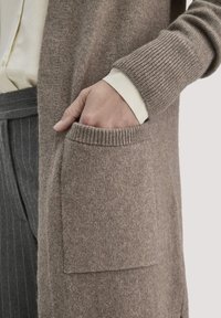 Mano nella tasca di un cardigan a maglia beige sopra il polsino di una camicia crema e pantaloni grigi a righe sottili, contro uno sfondo chiaro.