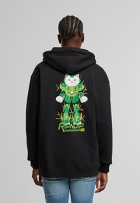 Persona con capelli intrecciati che indossa un hoodie nero con un gatto robotico verde con fulmini gialli e la scritta "RIPNDIP" sulla parte posteriore.