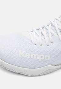 Bílé atletické boty s texturovaným síťovaným svrškem, zesílenou špičkou a bílými tkaničkami. Logo "Kempa" výrazně umístěné na boku.