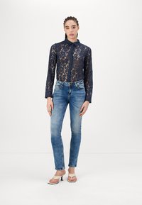 Chemise en dentelle bleu marine à manches longues et col, assortie à un jean skinny bleu clair et des talons beiges, présentant un design élégant et ajusté.