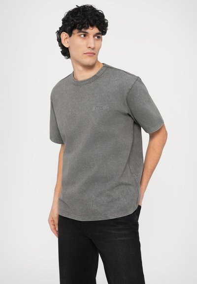 Szürke, rövid ujjú póló kerek nyakkivágással, puha anyaggal és finom logóval a mellrészen. Fekete farmerrel párosítva.