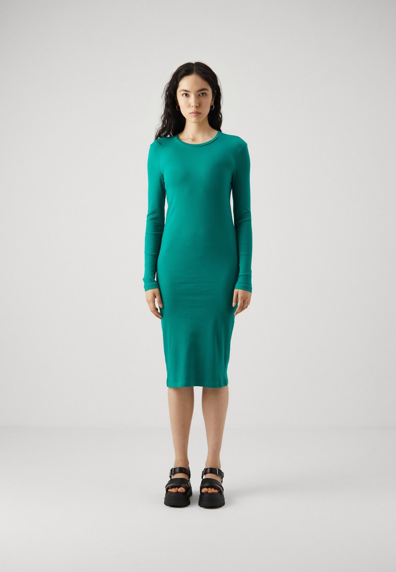 Τιρκουάζ φόρεμα με μακριά μανίκια, ριγωτή υφή και εφαρμοστή σιλουέτα, μέχρι το γόνατο, συνδυασμένο με χονδρές μαύρες σανδάλια. Απλή στρογγυλή λαιμόκοψη και χωρίς ραφές σχέδιο.