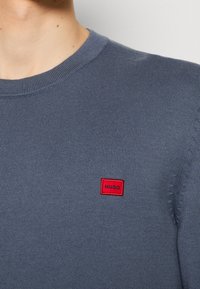 Gros plan d’une personne portant un pull en maille gris avec un petit patch rectangulaire rouge étiqueté "HUGO" sur la poitrine.