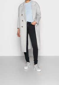 Abrigo largo gris claro con cuello de muesca, llevado sobre una camiseta azul claro y jeans ajustados negros. Zapatillas gris completan el conjunto.