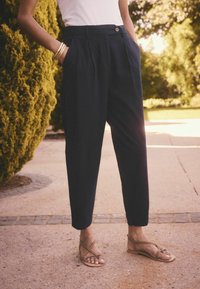 Marineblauwe broek met een losse pasvorm, geplooide voorkant en zijzakken, gecombineerd met tan sandalen. Omgeven door groen en asfalt.