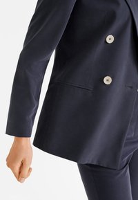 Tummanlaivansinen bleiseri, jossa on kaksirivinen malli, isot vaaleat napit ja sileä kangas, joka istuu hyvin.