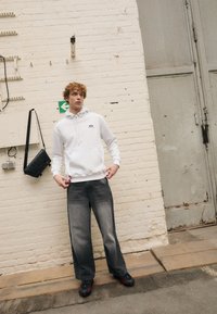 Witte capuchontrui met een voorzak en blauw logo, gecombineerd met donkergrijze wijde jeans en zwart-rode sneakers. Minimalistisch ontwerp.