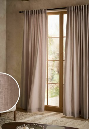 Tende beige chiaro appese a una finestra con cornice in legno, con un tappeto morbido e vista parziale di una sedia e un tavolino da caffè all'interno.