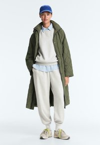 Person trägt langen, grünen Steppmantel, graue Jogginghosen, grauen Pullover über blau gestreiftem Hemd, blaue Mütze und beige Turnschuhe mit gelben Schnürsenkeln.