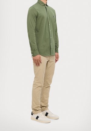Homme debout portant une chemise boutonnée vert olive, un pantalon beige et des baskets blanches avec des bandes noires sur fond blanc.