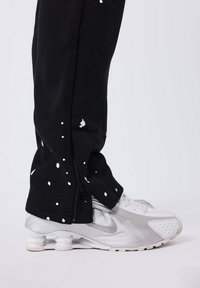 Zwarte sweatpants met witte spetters en ritsen bij de enkels, gecombineerd met zilveren en witte Nike sneakers met een kenmerkende hak.