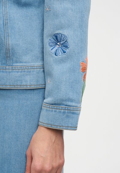Lyseblå denimjakkeærme med broderede blå og orange blomster over håndleddet, båret sammen med matchende denimbukser.