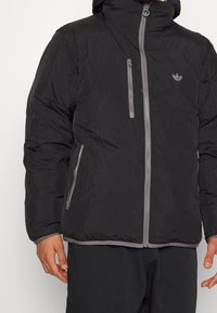 Svart puffjacka med huva, grå dragkedja och detaljer. Har två fickor med dragkedja på sidorna och en liten logotyp på bröstet.