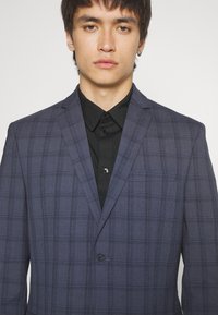Navy blue plaid blazer with notch lapels, single button, and a chest pocket, paired with a black collared shirt underneath.