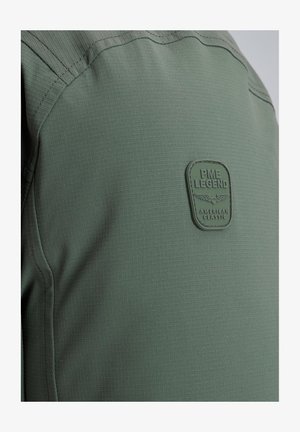 Vihreä kangaspaita, jossa on tekstuurinen ruutukuviokuvio ja näyttävä kuminen logo-osa, jossa lukee "PME Legend American Classic."