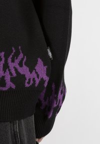 Svart stickad tröja med lila flamemönster på ärmarna och en liten etikett på sidan. Har ribbade manchetter och nederkant.