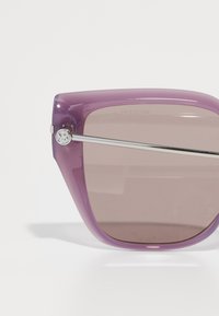 Ochelari de soare supradimensionați, de culoare mov, cu un finisaj lucios, lentile de culoare brună și accente metalice argintii pe brațe. Design al ramei unghiular.