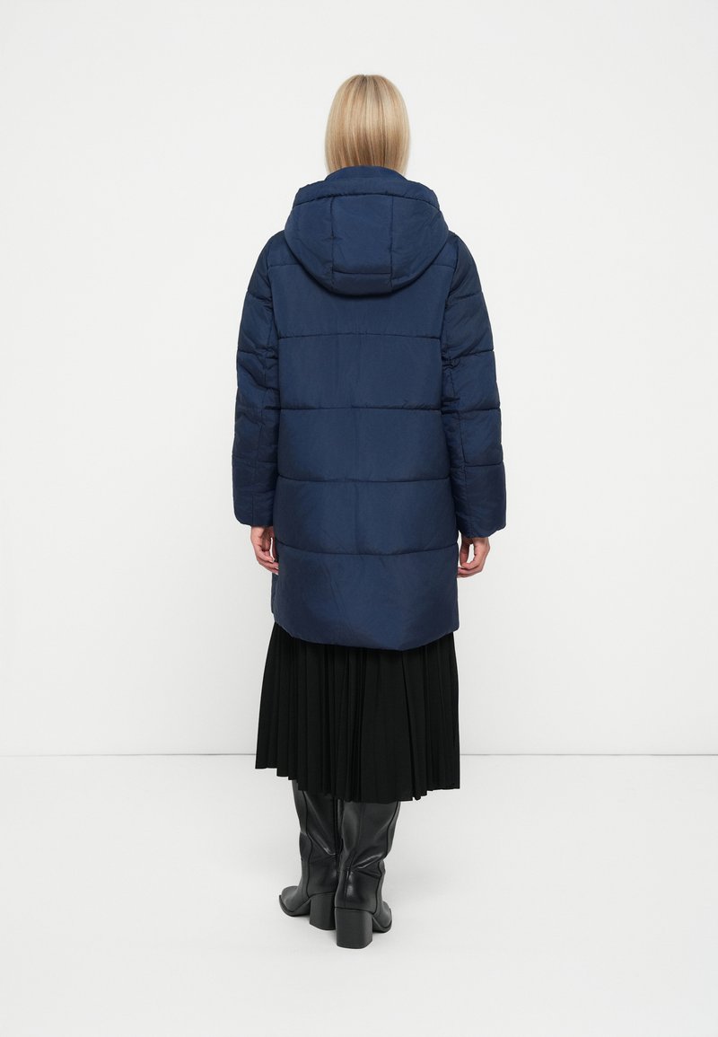 Cappotto puffo blu navy con cappuccio, design trapuntato e maniche lunghe, abbinato a una gonna plissettata nera e stivali neri fino al ginocchio.