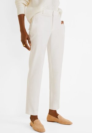 Personne à la peau foncée portant un pantalon sur mesure écru, un pull en maille crème et des mocassins en daim beige, debout sur un fond blanc.