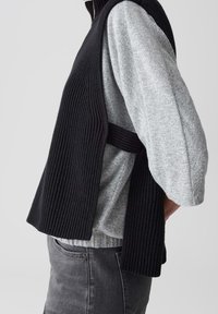 Vue latérale d'une personne portant un haut gris à manches longues et un gilet noir sans manches en maille côtelée avec des ouvertures latérales, sur un pantalon gris foncé.