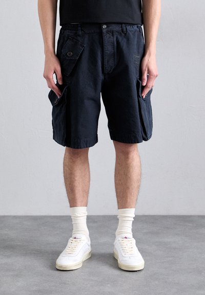 Shorts cargo bleu marine avec plusieurs grandes poches, fermeture à bouton et taille élastique ; associés avec des baskets blanches et des chaussettes de longueur mi-mollet.