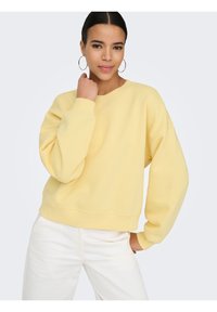 Νεαρή γυναίκα με κρίκους στα αυτιά που φοράει ένα φαρδύ ανοιχτό κίτρινο φούτερ και λευκό παντελόνι, ποζάροντας με το ένα χέρι κοντά στον λαιμό και το άλλο στην τσέπη.