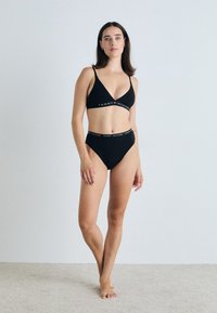 Fekete bikini szett, nyakba akasztható bralette-vel és magas derekú alsóval. Az anyag sima; a fejvédőn a "TOMMY HILFIGER" logó kontrasztos színekben látható.