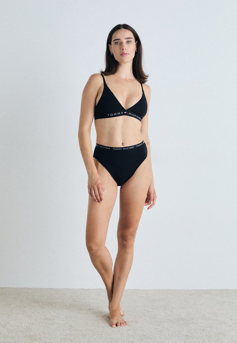 Črn bikini komplet, ki vključuje trikotno braletko in hlačke visokega pasu. Tkanina je gladka; pas ima v kontrastnih barvah napis "TOMMY HILFIGER".