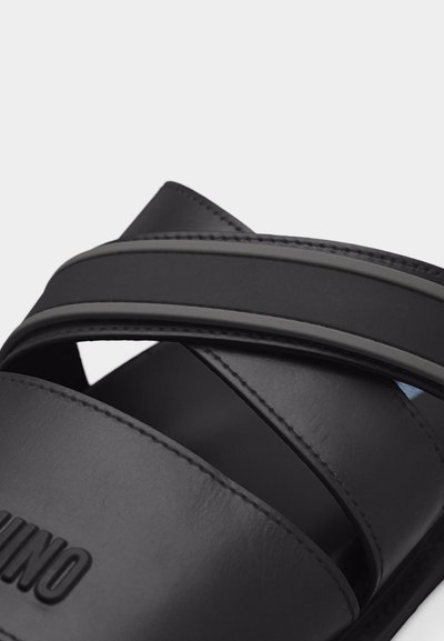 Gros plan sur une sandale noire à semelle plate avec deux larges brides, dont une avec un logo en relief.