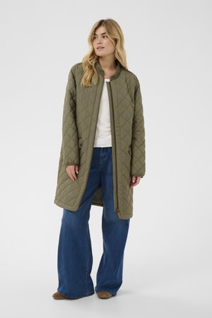 Nainen, jolla on pitkä oliivinvihreä toppatakki valkoisen paidan ja leveälahkeisten sinisten farkkujen päällä, seisoo yksiväristä taustaa vasten.