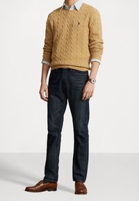 Sweter w splot kablowym w kolorze musztardowym, założony na jasnoniebieską koszulę z kołnierzykiem, ciemne dżinsy oraz brązowe skórzane buty. Prosty zegarek na nadgarstku, w pozycji stojącej.