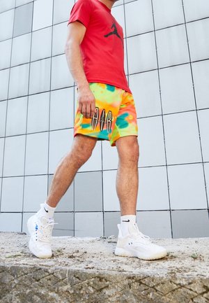 Camiseta roja con logo negro, pantalones cortos de teñido anudado multicolor en amarillo y verde con texto negro, y zapatos deportivos blancos con detalles en negro.