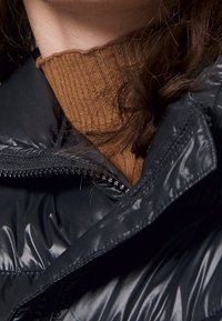 Black puffer jacket with shiny texture and exposed zipper, layered over a brown ribbed sweater. Details include a high collar and visible stitching.