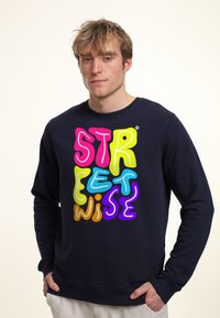 Námořnická mikina s barevným, vlnivým textem "STREETWISE", který obsahuje akcenty v růžové, zelené, modré, oranžové a fialové barvě. Klasický kulatý výstřih.