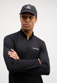 Juodos sportinės ilgomis rankovėmis marškinėliai su ketvirčio užtrauktuku ir lengva tekstūra, su balta Calvin Klein logo. Juoda kepurė su logotipu priekyje.