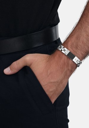 Bracciale in metallo con una combinazione di sezioni in argento lucido e nero testurizzato, indossato su un polso con una mano in tasca.