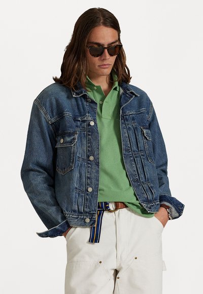 Zila džinsa jaka ar priekšējiem kabatām un sudrabainām pogām, virs zaļas polo krekliņa. Modelis nēsā saulesbrilles un baltas bikses ar jostu.