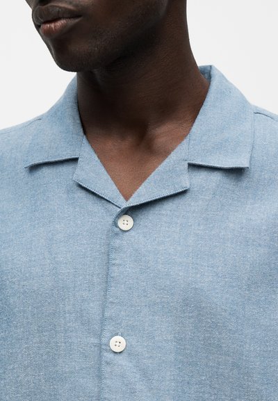 Gros plan d'une personne portant une chemise bleu clair à col avec deux boutons blancs attachés à l'avant.