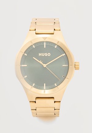 Ceas de mână auriu cu cadran verde, marcaje orare minimaliste, trei ace arătând ora 10:09:21 și logo-ul "HUGO" în centrul cadranului.