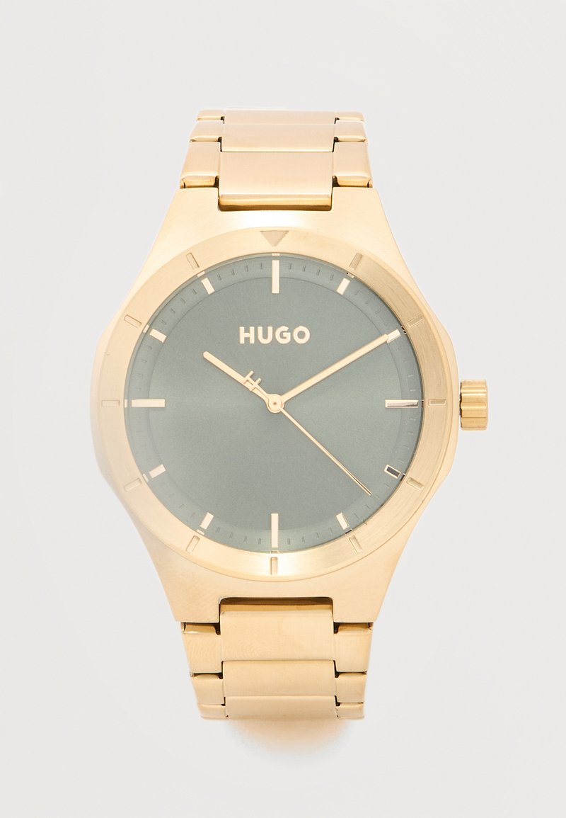 Χρυσό ρολόι χειρός με πράσινο καντράν, ελάχιστους δείκτες ώρας, τρεις δείκτες που δείχνουν 10:09:21 και λογότυπο "HUGO" στο κέντρο.
