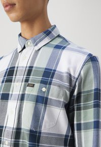 Gros plan d'une personne portant une chemise à carreaux bleue, verte et blanche avec des boutons et une petite poche poitrine étiquetée Lee.