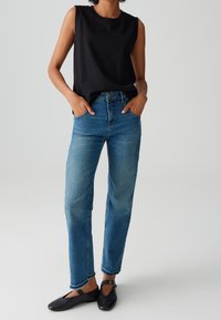 Schwarzes ärmelloses Oberteil, blaue hochtaillierte gerade Jeans und schwarze flache Schuhe mit Riemen. Glatte Textur, lässiges Design.