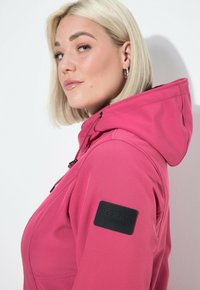 Růžová kapucňová bunda s texturovanou tkaninou, s černou nášivkou loga na levém rukávu a stahovací šňůrkou pro nastavitelný střih.