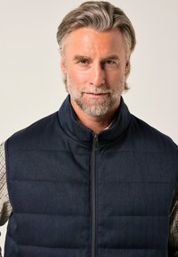 Mörkblå puffväst med hög krage och fiskbensmönster, med en frontdragkedja och quiltat mönster. Bärs över en rutmönstrad skjorta.