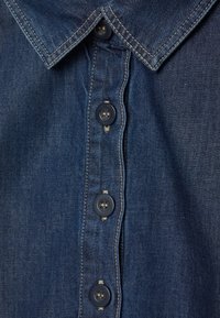 Detailní záběr na límec modré denimové košile a tři tmavě námořnické knoflíky s béžovým šitím podél légy knoflíků.