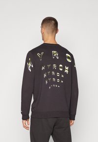 Svart sweatshirt laget av bomull med lange ermer, med hevet, flerfargede "HYROX"-grafikker på ryggen og ermene.